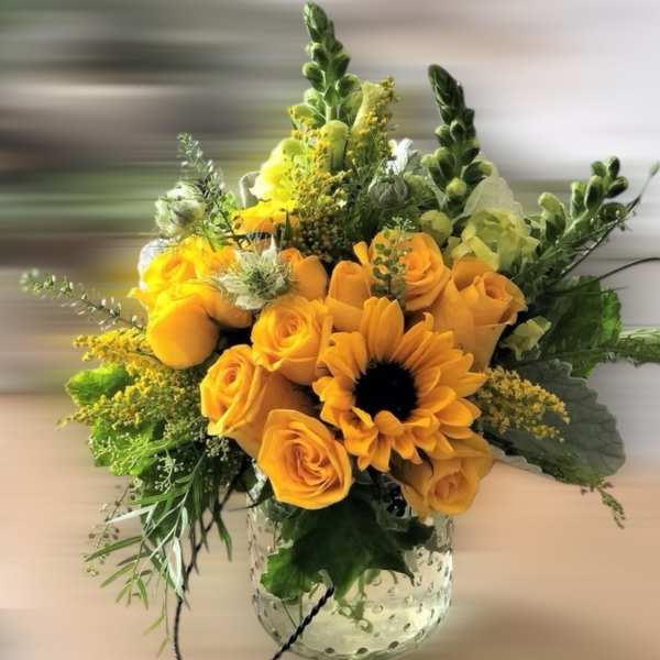 Yellow roses and a sunflower arranged with accent blooms in a clear textured glass vase