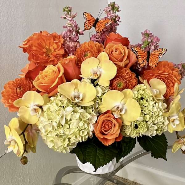 Orange and yellow floral arrangement with roses, orchids, and butterflies in a white container