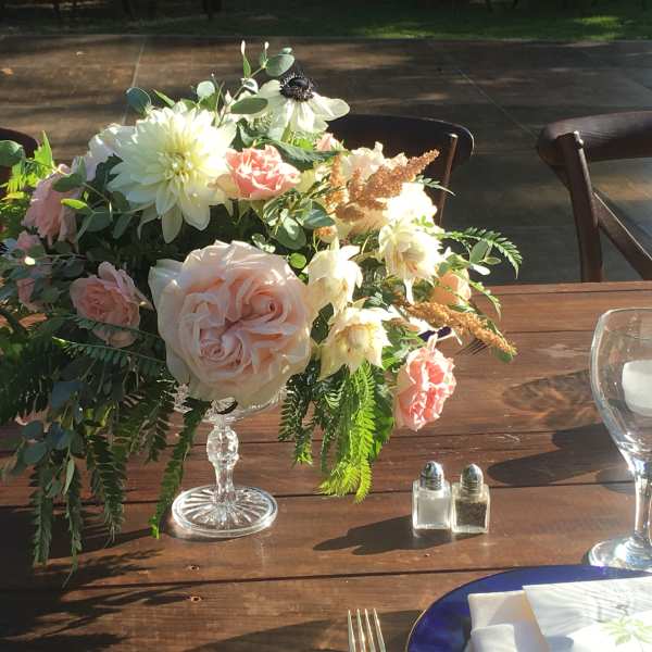 Pastel floral centerpiece in a clear pedestal vase on a wooden table