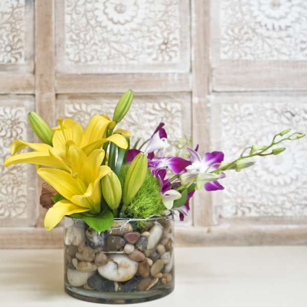 Yellow lilies and purple orchids in a glass vase filled with pebbles