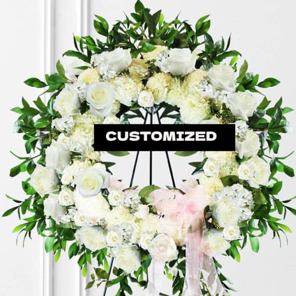 White floral wreath on a stand with green foliage and a pink ribbon