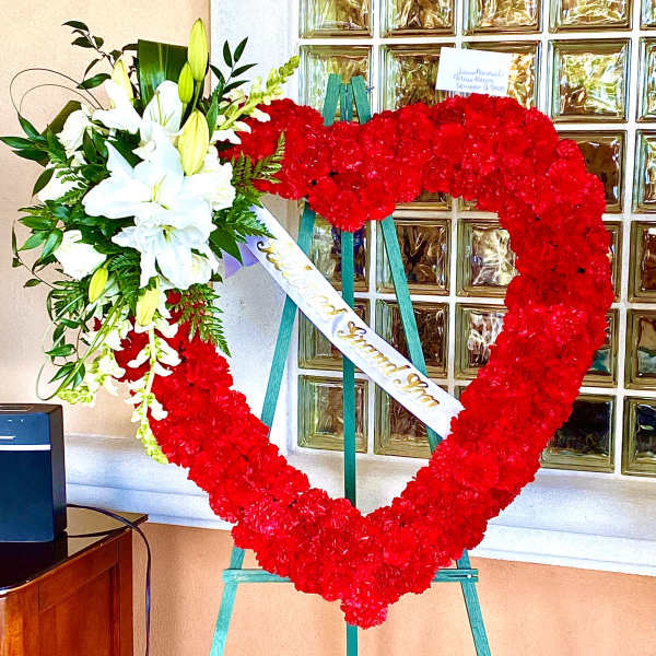 Heart-shaped red floral wreath on an easel with white lilies and a ribbon