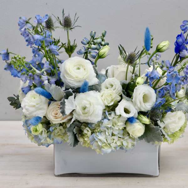 Low arrangement of white roses and ranunculus with blue delphinium in a gray ceramic vase