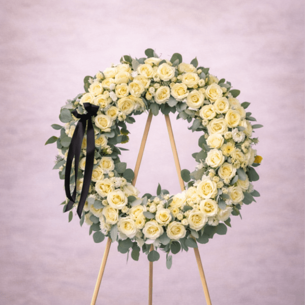 Large round wreath of pale yellow roses on a wooden easel with a black ribbon.