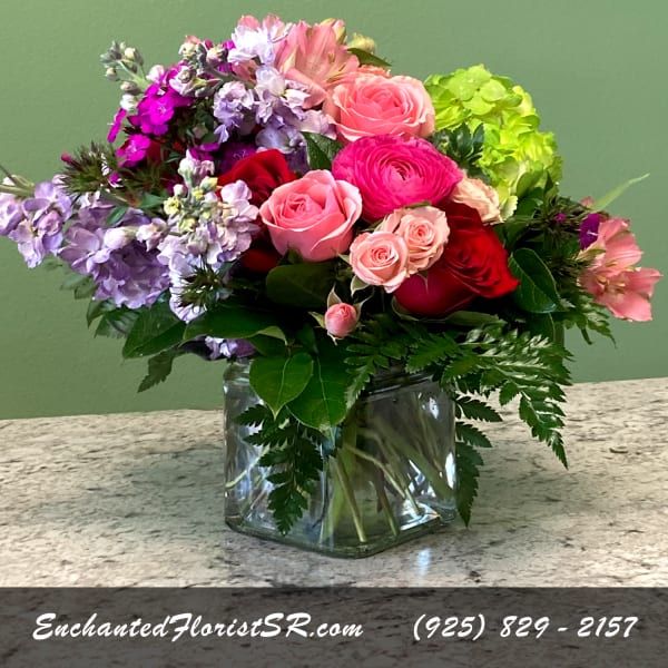 Mixed arrangement of pink and red roses with purple flowers in a clear glass cube vase