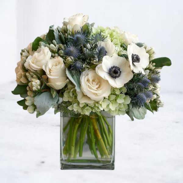 Low glass vase of white roses, white anemones, green hydrangeas and blue accent blooms