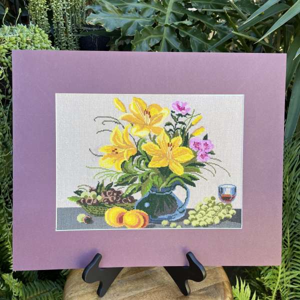Framed floral needlepoint with yellow lilies and fruit on a stand