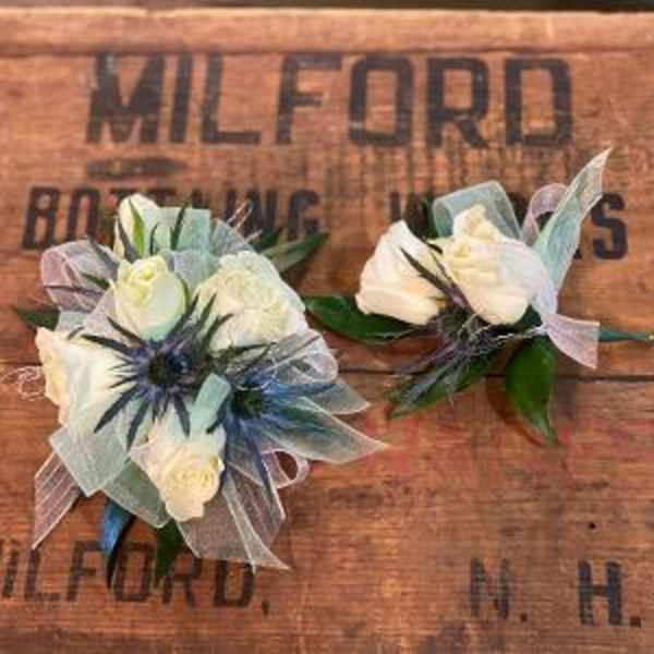 Two white floral boutonnieres with blue accents on a wooden surface