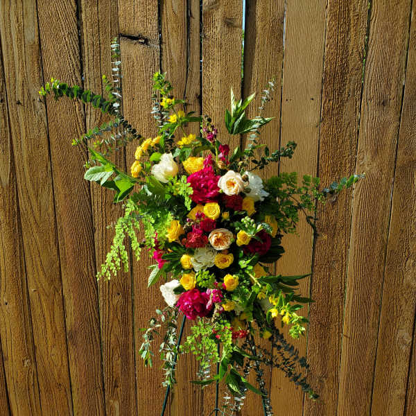Bright standing spray of yellow, red, and white flowers on a metal easel against a wood fence.