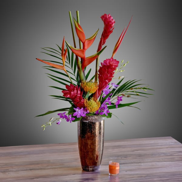 Tropical flower arrangement in a tall metallic vase with a small candle nearby