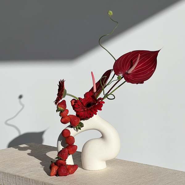 Red anthuriums and gerbera daisies in a sculptural white vase with strawberries draped down the side.