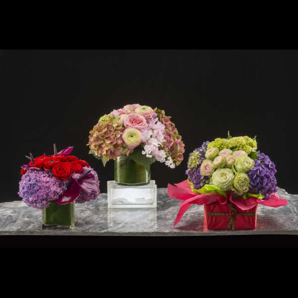 Three colorful floral arrangements in glass and gift boxes on a marble surface
