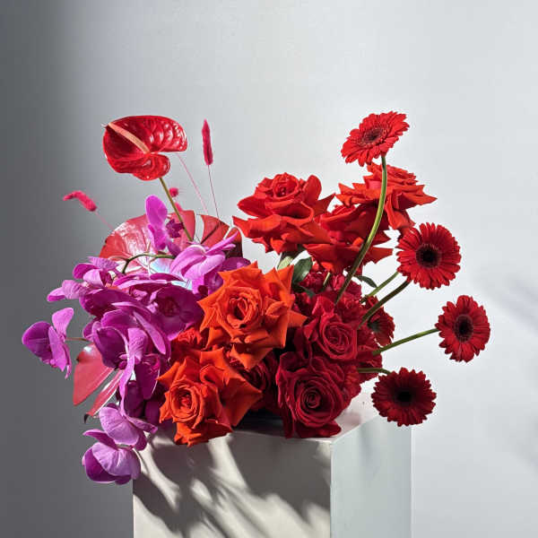 Red and purple floral arrangement with roses and gerbera daisies in a white pedestal vase