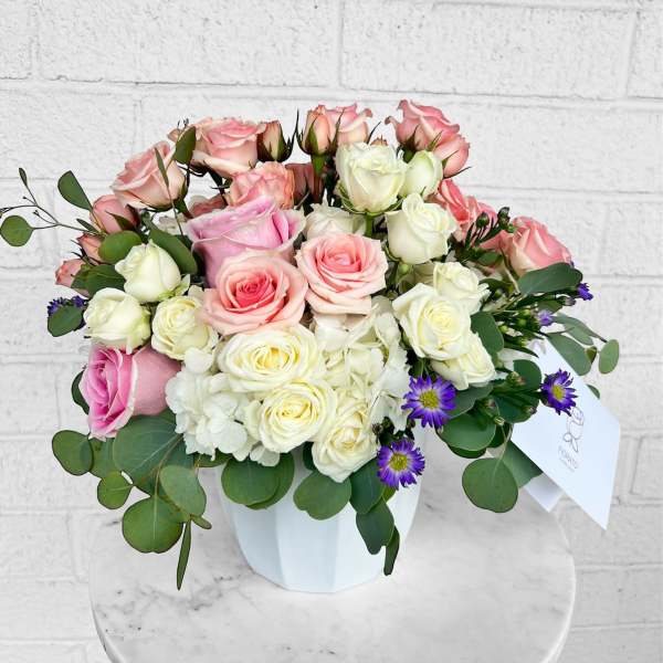 Pink and white roses arranged in a white box with purple accent flowers.