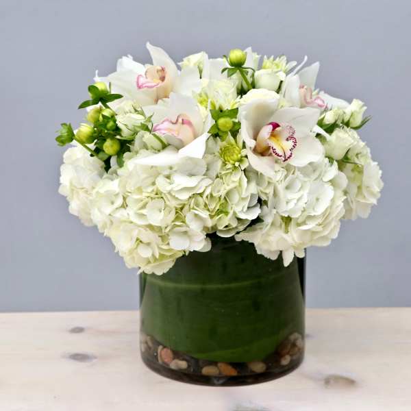 Low round arrangement of white hydrangeas and orchids in a glass cylinder vase with stones.