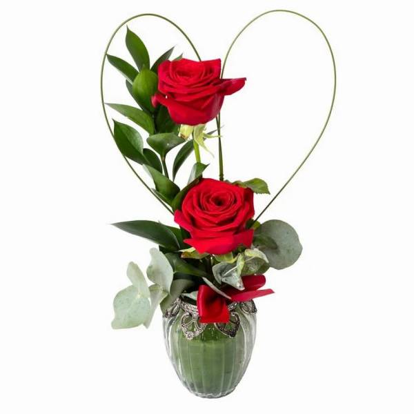 Two red roses in a glass vase with a heart-shaped wire frame