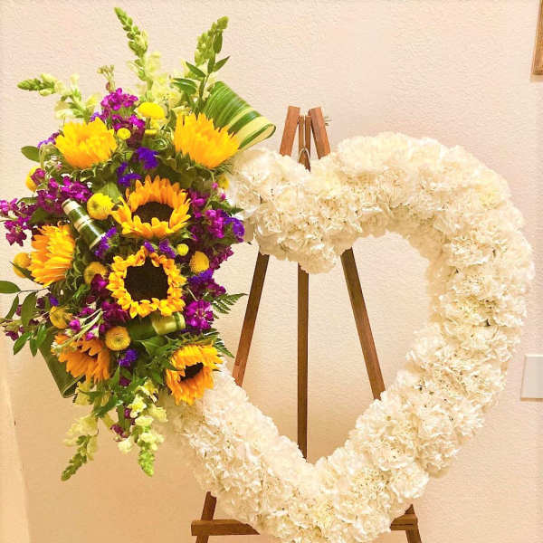 Heart-shaped white floral wreath on an easel beside a colorful sunflower bouquet.