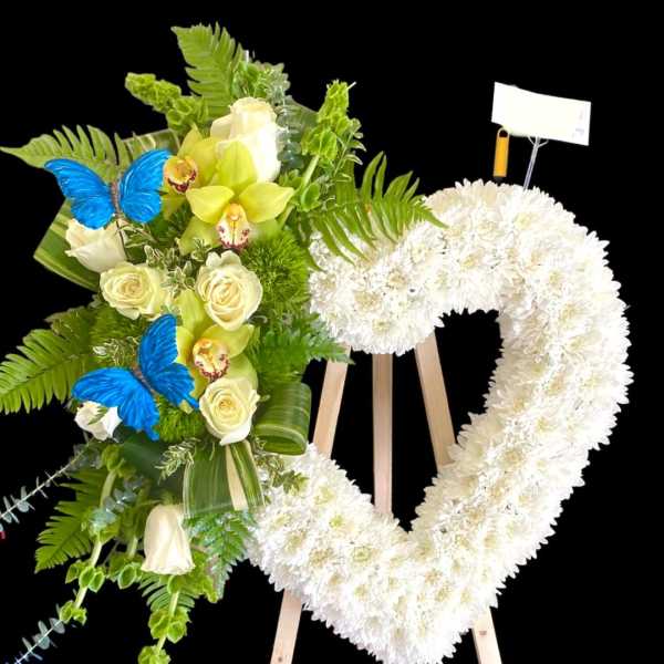 Heart-shaped white floral wreath on an easel beside a green and white bouquet with blue butterflies
