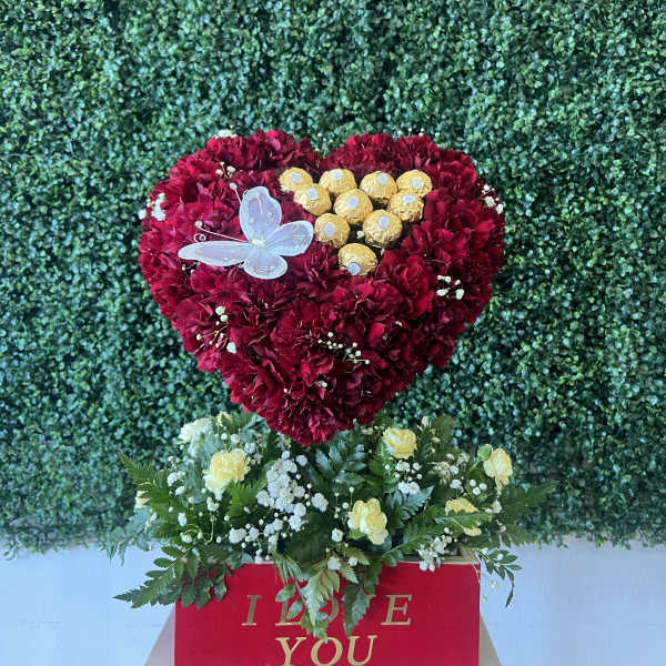Heart-shaped red carnation arrangement with chocolates and a butterfly topper