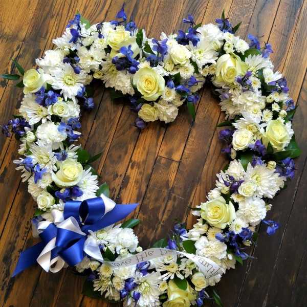 Heart-shaped wreath of white flowers with yellow roses, blue blooms, and a blue and white ribbon bow