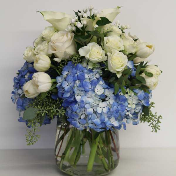 Blue hydrangeas with white roses, tulips, and calla lilies in a clear glass vase