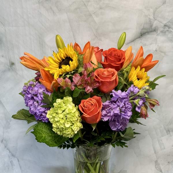 Mixed bouquet of roses, sunflowers, lilies, and purple blooms in a glass vase