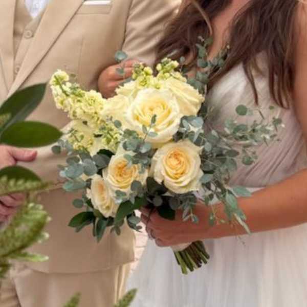 Bridal hand-tied bouquet of pale yellow roses and cream flowers with soft greenery, held by a bride in white.