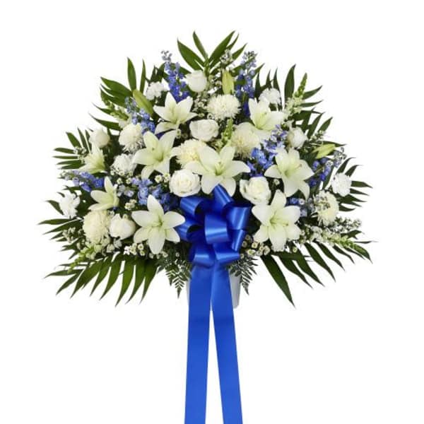Standing spray of white lilies, roses, and carnations with blue flowers and a blue ribbon bow