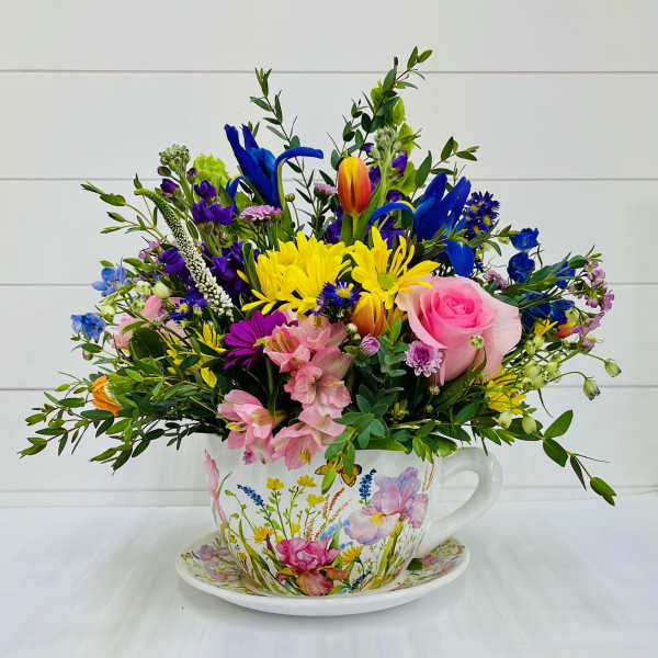 Colorful mixed flower arrangement in a floral teacup and saucer with pink, yellow, blue and purple blooms