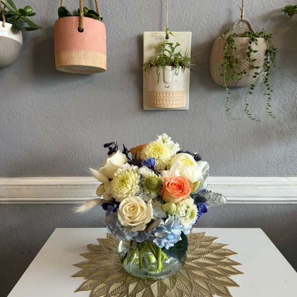 Mixed bouquet in a clear glass vase with cream, peach, blue, and yellow flowers.