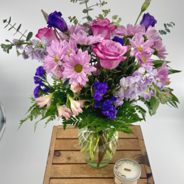 Mixed bouquet of pink roses and purple blooms in a glass vase on a wooden crate with a small candle.