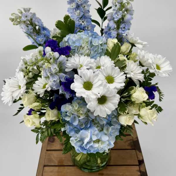 Blue hydrangeas, white daisies, and cream roses arranged in a clear glass vase.