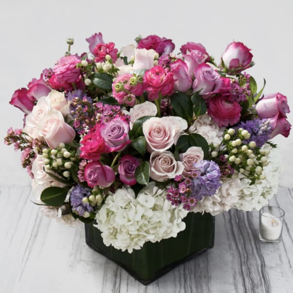Low arrangement of pink and lavender roses with white hydrangeas in a green-wrapped glass vase.