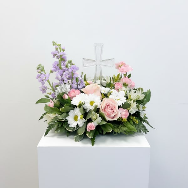 Low pastel arrangement of pink roses, white daisies, and lavender blooms with a clear glass cross in the center