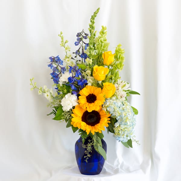 Tall mixed bouquet in a blue vase with sunflowers, roses, delphinium, and hydrangea
