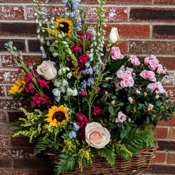 Tall mixed arrangement with sunflowers, roses, blue delphinium, and pink flowering plant in a wicker basket.