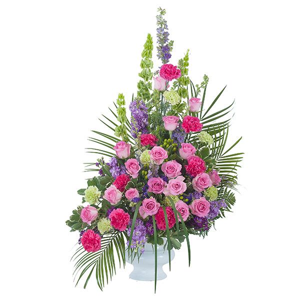 Tall pink and purple arrangement with roses and carnations in a white urn accented by palm fronds.