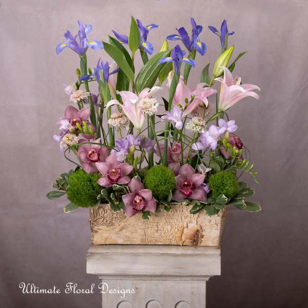 Tall floral arrangement with pink lilies, purple orchids, and green pom-pom blooms in a wooden box