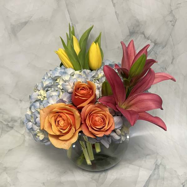 Bouquet of orange roses, yellow tulips, blue hydrangea, and pink lilies in a glass vase