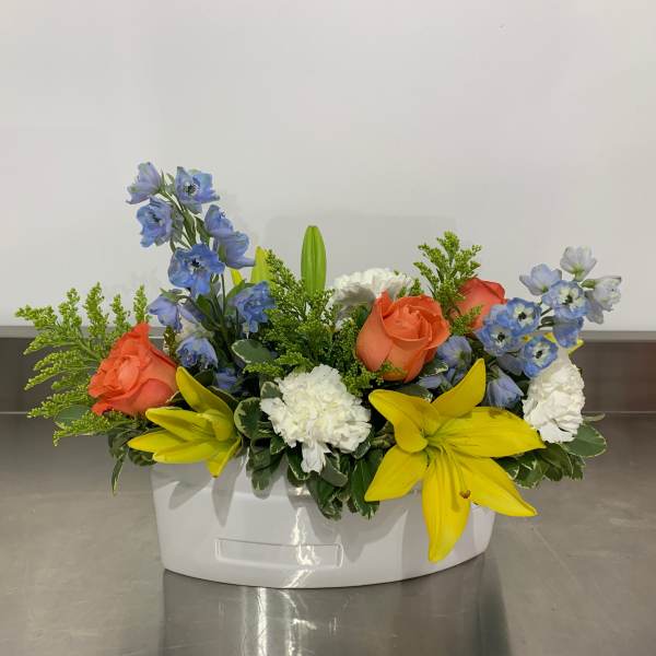 Mixed floral arrangement in a white container with orange roses, blue flowers, and yellow lilies
