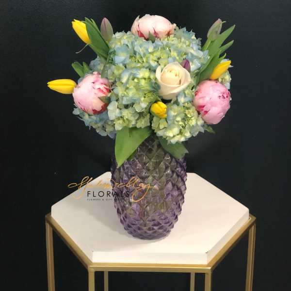 Mixed bouquet in a purple glass vase with blue hydrangeas, pink peonies, and yellow tulips