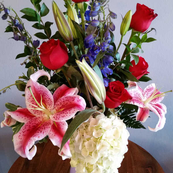 Mixed bouquet with red roses, pink lilies, blue delphinium, and white hydrangea in a lavender vase