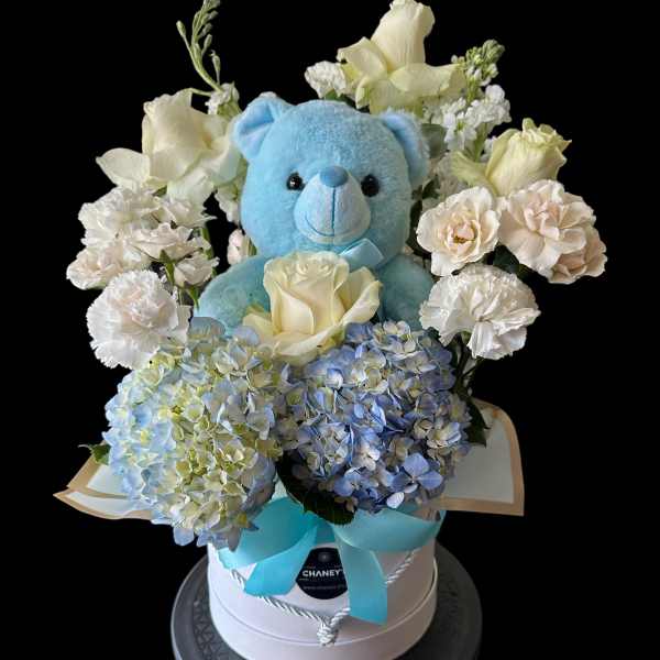 Blue teddy bear surrounded by white roses and blue hydrangeas in a gift box