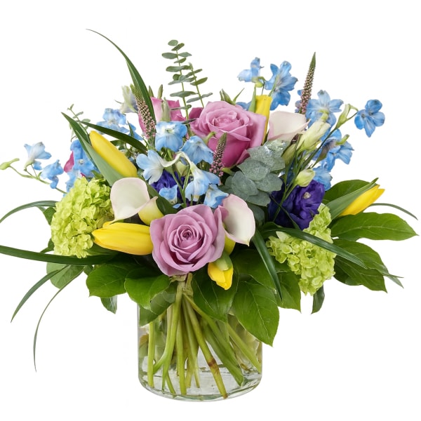 Mixed bouquet of pink roses, blue flowers, yellow tulips, and white calla lilies in a glass vase