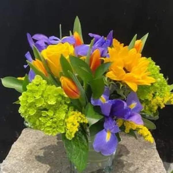 Bright arrangement of yellow flowers, purple irises, and orange tulips in a clear glass cube vase