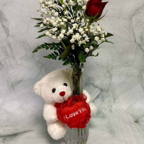 Two red roses in a glass vase with a teddy bear holding a heart