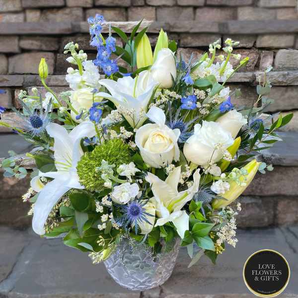 White lilies and roses with blue accents arranged in a clear patterned glass vase