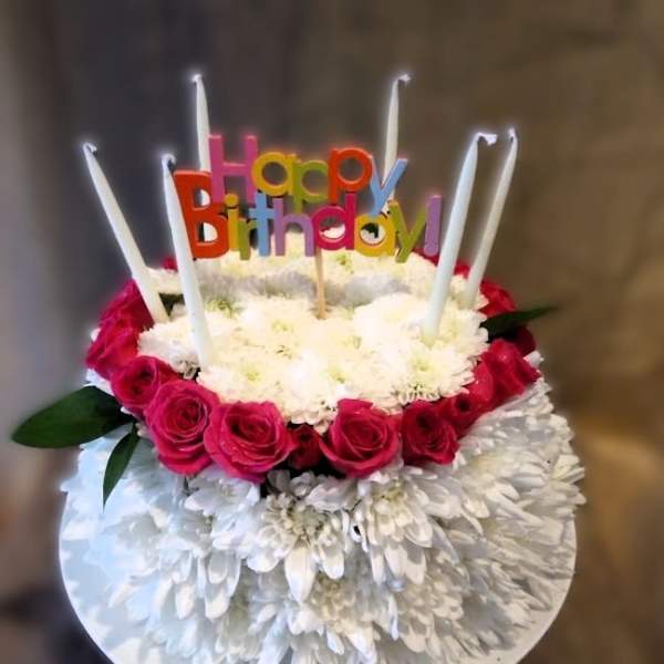 Round cake-shaped arrangement of white mums and pink roses with tall candles and a Happy Birthday pick