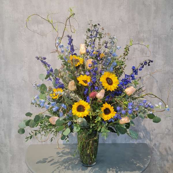 Tall bouquet of sunflowers, blue flowers, and peach roses in a glass vase