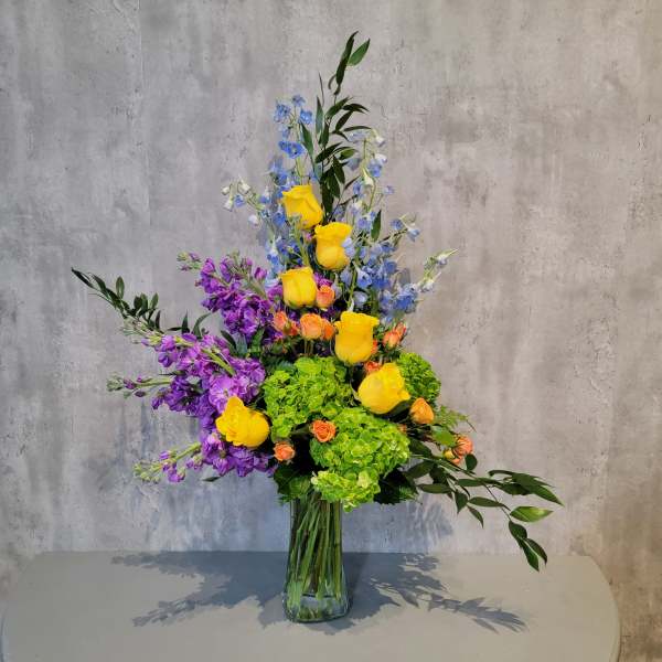 Tall mixed bouquet in a clear glass vase with yellow, purple, blue, and green blooms